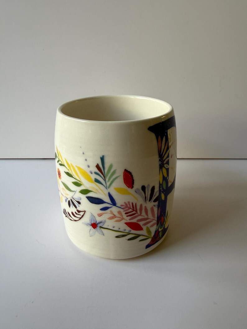 May include: A cream-colored ceramic mug with a floral design. The mug features a colorful illustration of leaves, flowers, and branches in shades of blue, yellow, red, and pink. The mug has a cylindrical shape and a smooth surface.