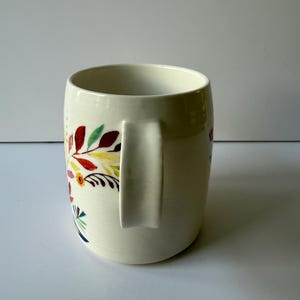 May include: Cream-colored ceramic mug with a curved handle. The mug is decorated with a colorful floral pattern featuring red, yellow, green, and blue leaves and flowers. The mug has a wide opening and a smooth, glossy finish.