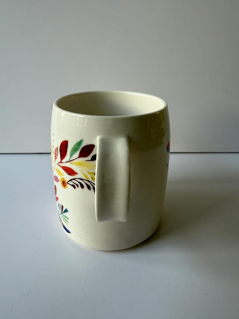 May include: Cream-colored ceramic mug with a curved handle. The mug is decorated with a colorful floral pattern featuring red, yellow, green, and blue leaves and flowers. The mug has a wide opening and a smooth, glossy finish.