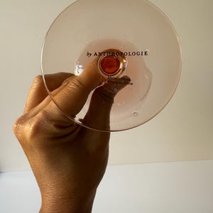May include: A pink-tinted glass with a round bowl and a stem, held by a hand. The text "by ANTHROPOLOGIE" is printed on the glass. The glass is made in China.