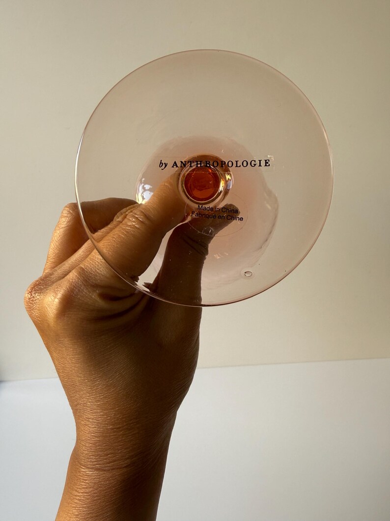 May include: A pink-tinted glass with a round bowl and a stem, held by a hand. The text "by ANTHROPOLOGIE" is printed on the glass. The glass is made in China.