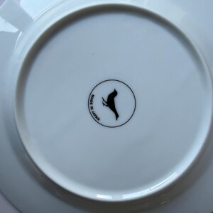 May include: A white ceramic plate with a black bird logo and the text "MADE IN JAPAN" in a circular design. The plate has a slightly raised edge and a smooth, glossy surface.