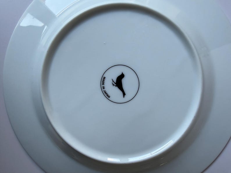 May include: A white ceramic plate with a black bird logo and the text "MADE IN JAPAN" in a circular design. The plate has a slightly raised edge and a smooth, glossy surface.