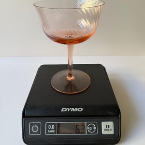 May include: A pink-tinted glass champagne coupe with a spiral design sits on a black digital scale. The scale displays a weight of 6.3 lbs. The coupe has a rounded bowl and a slender stem.