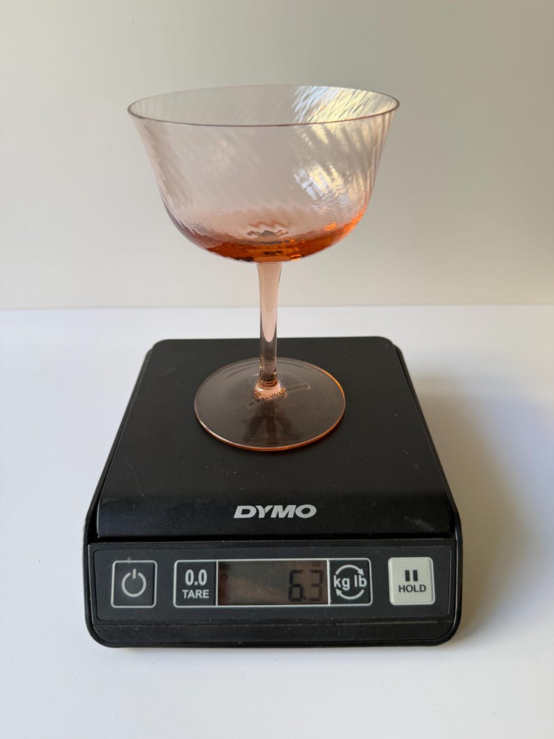 May include: A pink-tinted glass champagne coupe with a spiral design sits on a black digital scale. The scale displays a weight of 6.3 lbs. The coupe has a rounded bowl and a slender stem.