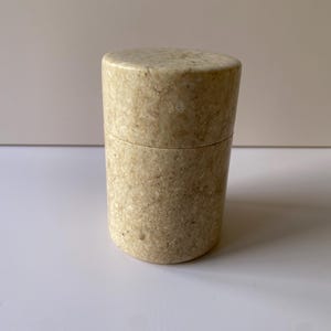 May include: A cylindrical container with a lid. The container is made of a speckled, beige material. The lid sits flush with the body of the container. The container is on a white surface.