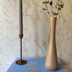 Op de afbeelding: Een blauw boek met de titel "HUE KELLY WEARSTLER" op de omslag. Een gouden kandelaar met een grijze kaars en een getextureerde, lichtroze vaas met groene bladeren staan op het boek.