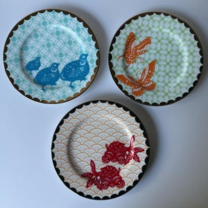 May include: Three decorative plates with unique designs. One plate features blue birds on a turquoise background, another has orange butterflies on green, and the last showcases red goldfish on a cream background. Each plate has a scalloped edge with a gold or black trim.