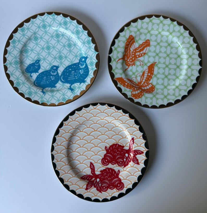 May include: Three decorative plates with unique designs. One plate features blue birds on a turquoise background, another has orange butterflies on green, and the last showcases red goldfish on a cream background. Each plate has a scalloped edge with a gold or black trim.