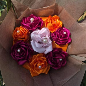 Peut inclure: Un bouquet de roses en ruban faites à la main, dans des tons d'orange, de fuchsia et de rose, chacune ornée d'une perle en son centre. L'arrangement est présenté dans un emballage en papier brun, créant une esthétique artisanale charmante.