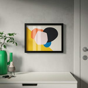 Peut inclure: Une impression d'art encadrée avec des formes géométriques orange, rose, noir et bleu sur un fond jaune et blanc. L'œuvre est dans un cadre noir, accrochée à un mur gris clair au-dessus d'une commode blanche.