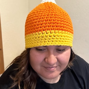 May include: A crocheted candy corn hat with a white tip, orange body, and yellow bottom. The hat is worn by a person with brown hair and a black shirt with the text "The Forgers" and "Void Forgers" printed on it.