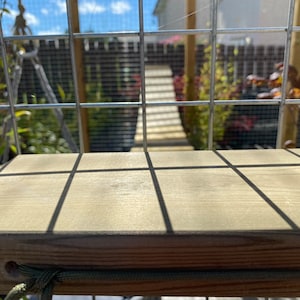 Könnte beinhalten: Ein hellbraunes Katzenregal aus Holz, rechteckig, an einer Drahtgitterstruktur befestigt. Das Regal hat ein Seildetail. Der Hintergrund zeigt einen Steg und Pflanzen, mit Schatten der Sonne, die auf das Regal fallen.