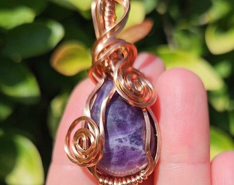May include: A purple amethyst gemstone wrapped in copper wire, creating a teardrop-shaped pendant.
