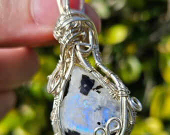 May include: A silver wire wrapped pendant featuring a rainbow moonstone with black inclusions. The stone is set in a teardrop shape and the wire wrapping creates a delicate and intricate design.