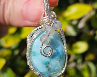 May include: A handcrafted pendant featuring a light blue larimar stone, wire-wrapped in silver. The oval-shaped stone has a swirl design and is accented with small, round, gray beads. The pendant is suspended from a silver wire loop.