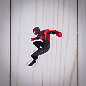 May include: A sticker featuring a stylized illustration of a superhero in a dynamic pose. The character is depicted in a black suit with red accents on the arms and chest. The superhero is jumping with a clenched fist.