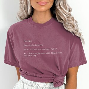 May include: A maroon t-shirt with a white graphic that defines the word "boujee" as an adjective meaning "rich, luxurious, special, fancy". The graphic also includes an example sentence: "She's so boujee with that Louis Vuitton bag."
