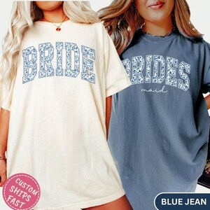 May include: Two t-shirts, one cream-colored and one blue, with floral text. The cream shirt says "BRIDE" in blue floral letters. The blue shirt says "BRIDESMAID" in blue floral letters with "maid" in script below.