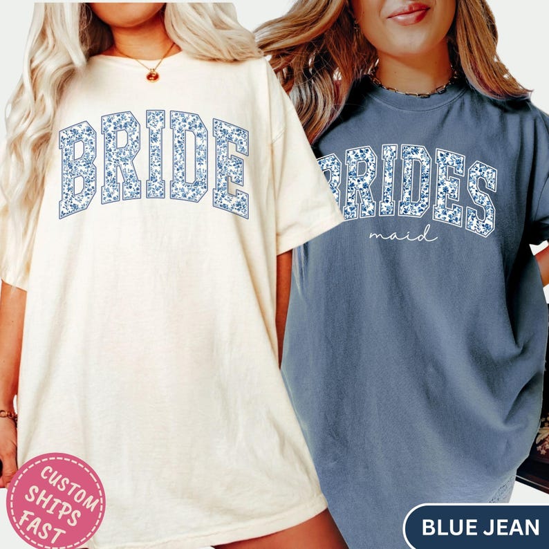 May include: Two t-shirts, one cream-colored and one blue, with floral text. The cream shirt says "BRIDE" in blue floral letters. The blue shirt says "BRIDESMAID" in blue floral letters with "maid" in script below.