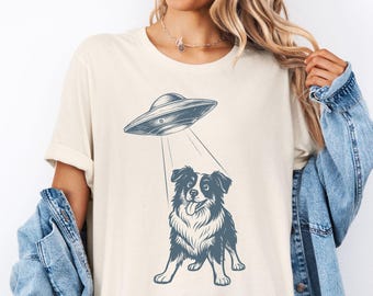 May include: Cream-colored t-shirt featuring a blue illustration of a UFO beaming a dog. The dog has black and white fur and is sticking out its tongue. The shirt has rolled-up sleeves.