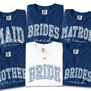May include: A collection of navy blue and white t-shirts with floral text designs. The shirts feature phrases like "BRIDE", "MAID of honor", and "MOTHER of the bride". One white shirt is also included.