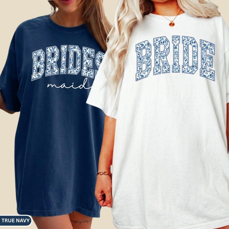 May include: Two oversized t-shirts, one navy blue and one white. The navy shirt says "BRIDESMAID" in white floral print, and the white shirt says "BRIDE" in blue floral print. Both shirts have short sleeves and a relaxed fit.