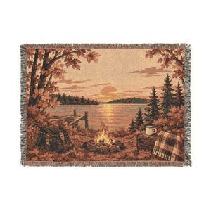 May include: A woven throw blanket featuring a landscape scene of a lake at sunset with a campfire in the foreground. The image includes a wooden dock, pine trees, a backpack, a mug, and a plaid blanket, all rendered in warm autumn tones.