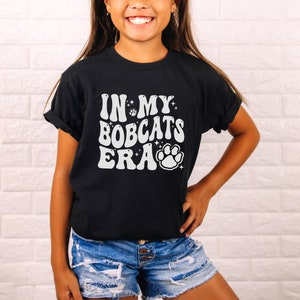 K&ouml;nnte beinhalten: Ein schwarzes T-Shirt mit einem wei&szlig;en Aufdruck, der "In My Bobcats Era" mit einem Pfotenabdruck und Sternen zeigt.