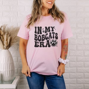 K&ouml;nnte beinhalten: Ein pinkfarbenes T-Shirt mit dem Text "In My Bobcats Era" in Schwarz mit einem Pfotenabdruck und Sternen.