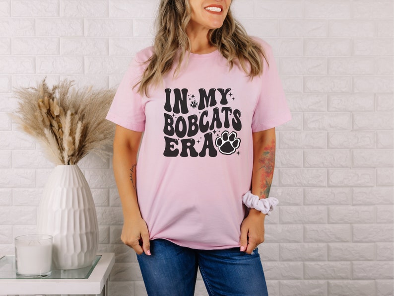 K&ouml;nnte beinhalten: Ein pinkfarbenes T-Shirt mit dem Text "In My Bobcats Era" in Schwarz mit einem Pfotenabdruck und Sternen.