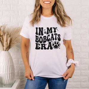 K&ouml;nnte beinhalten: Wei&szlig;es T-Shirt mit schwarzem Text, der "In My Bobcats Era" lautet, mit einem Pfotenabdruck-Motiv.