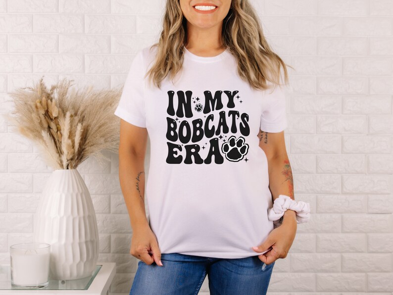 K&ouml;nnte beinhalten: Wei&szlig;es T-Shirt mit schwarzem Text, der "In My Bobcats Era" lautet, mit einem Pfotenabdruck-Motiv.