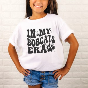 K&ouml;nnte beinhalten: Wei&szlig;es T-Shirt mit schwarzem Text, der "In My Bobcats Era" lautet, mit einem Pfotenabdruck-Motiv.