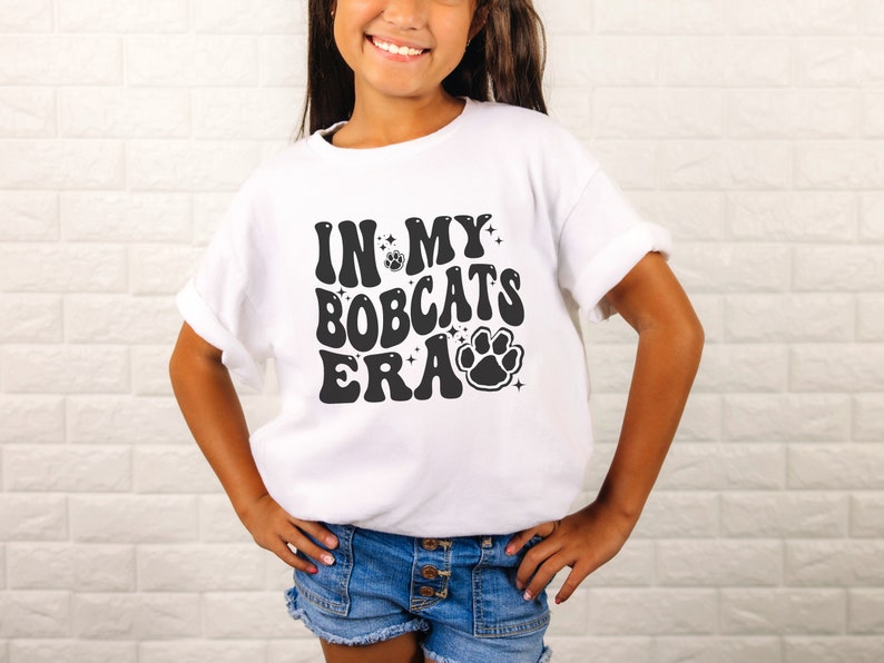 K&ouml;nnte beinhalten: Wei&szlig;es T-Shirt mit schwarzem Text, der "In My Bobcats Era" lautet, mit einem Pfotenabdruck-Motiv.
