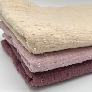 Könnte beinhalten: Ein Stapel von vier gefalteten Tüchern in verschiedenen Rosa- und Beigetönen. Das oberste Tuch ist hellbeige, gefolgt von einem blassen Rosa, einem mittleren Rosa und einem tiefen Malve. Jedes Tuch hat ein strukturiertes, gepunktetes Muster.