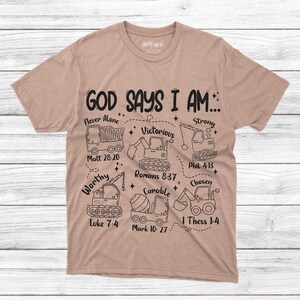 Puede incluir: Una camiseta color beige con la frase "GOD SAYS I AM..." y palabras inspiradoras con referencias b&iacute;blicas, como "Never Alone" y "Victorious". El dise&ntilde;o incluye dibujos lineales de veh&iacute;culos de construcci&oacute;n. La camiseta est&aacute; hecha de un material suave.