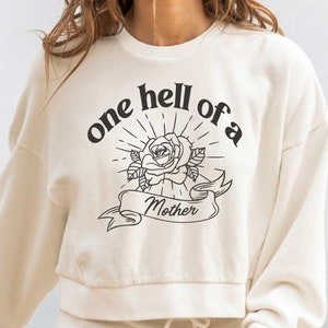 May include: A white sweatshirt with the text "one hell of a" in a black circular design with a rose and banner that reads "Mother" in the center.