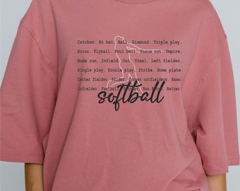 May include: A pink t-shirt with a white silhouette of a softball player and the text "Catcher. At bat. Ball. Diamond. Triple play. Error. Flyball. Foul ball. Force out. Umpire. Home run. Infield. Out. Steal. Left fielder. Single play. Double play. Strike. Home plate. Center fielder. Slider. Corner outfielders. Base. Infielder. Fastball Pitcher. Run. Batter. softball"