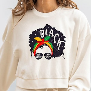 Puede incluir: Sudadera blanca con una ilustración en blanco y negro de una mujer con afro que lleva una diadema roja, amarilla y verde y gafas de sol. El texto "BLACK" está escrito en una fuente estilizada sobre la cabeza de la mujer. Las gafas de sol tienen el texto "NO SUGAR" y "NO CREAM" escrito en ellas.