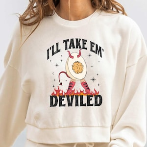 May include: A white cropped sweatshirt with a graphic of a deviled egg with horns and red shoes. The text "I'll take 'em deviled" is printed on the sweatshirt.