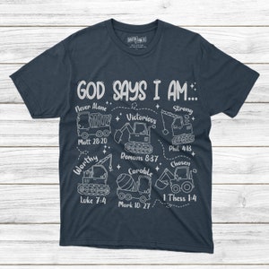 Puede incluir: Camiseta azul marino con texto blanco e ilustraciones de veh&iacute;culos de construcci&oacute;n. El texto dice "GOD SAYS I AM..." con frases como "Never Alone" y "Victorious" y vers&iacute;culos b&iacute;blicos. Un dise&ntilde;o &uacute;nico e inspirador.