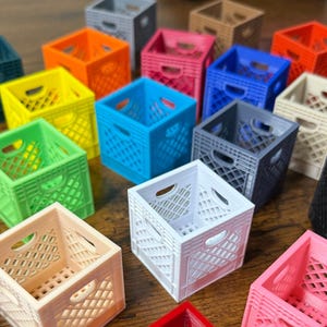 May include: A collection of miniature milk crates in various colours, including green, blue, yellow, orange, pink, white, grey, and black. The crates are made of plastic and have a grid pattern.