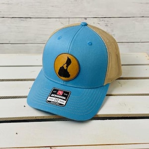 May include: A light blue trucker hat with a brown leather patch featuring the outline of an island. The hat has a mesh back and a Richardson 112 sticker.