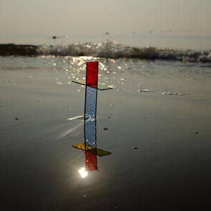 May include: A cross-shaped stained glass ornament featuring a vertical piece with a textured clear glass base and a red top section, intersected by a horizontal green glass bar. The object stands upright, casting a clear reflection on the wet surface.