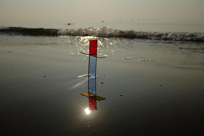 May include: A cross-shaped stained glass ornament featuring a vertical piece with a textured clear glass base and a red top section, intersected by a horizontal green glass bar. The object stands upright, casting a clear reflection on the wet surface.