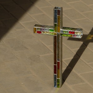 May include: A standing cross constructed from multiple small, rectangular pieces of stained glass in shades of red, green, yellow, and clear, held together by thin metal soldering. The cross features a grid-like pattern of colored glass segments.