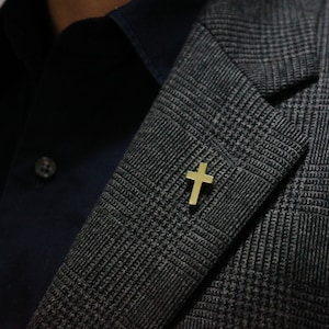 Könnte beinhalten: Eine goldfarbene Kreuz-Reversnadel auf einem grauen gemusterten Blazer.