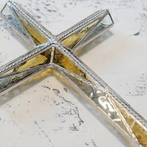 May include: A decorative cross featuring a central panel of textured amber-colored glass encased in a polished silver-toned metal frame. The frame has a twisted rope-like border detail along the edges, creating a structured and reflective appearance.