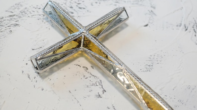 May include: A decorative cross featuring a central panel of textured amber-colored glass encased in a polished silver-toned metal frame. The frame has a twisted rope-like border detail along the edges, creating a structured and reflective appearance.